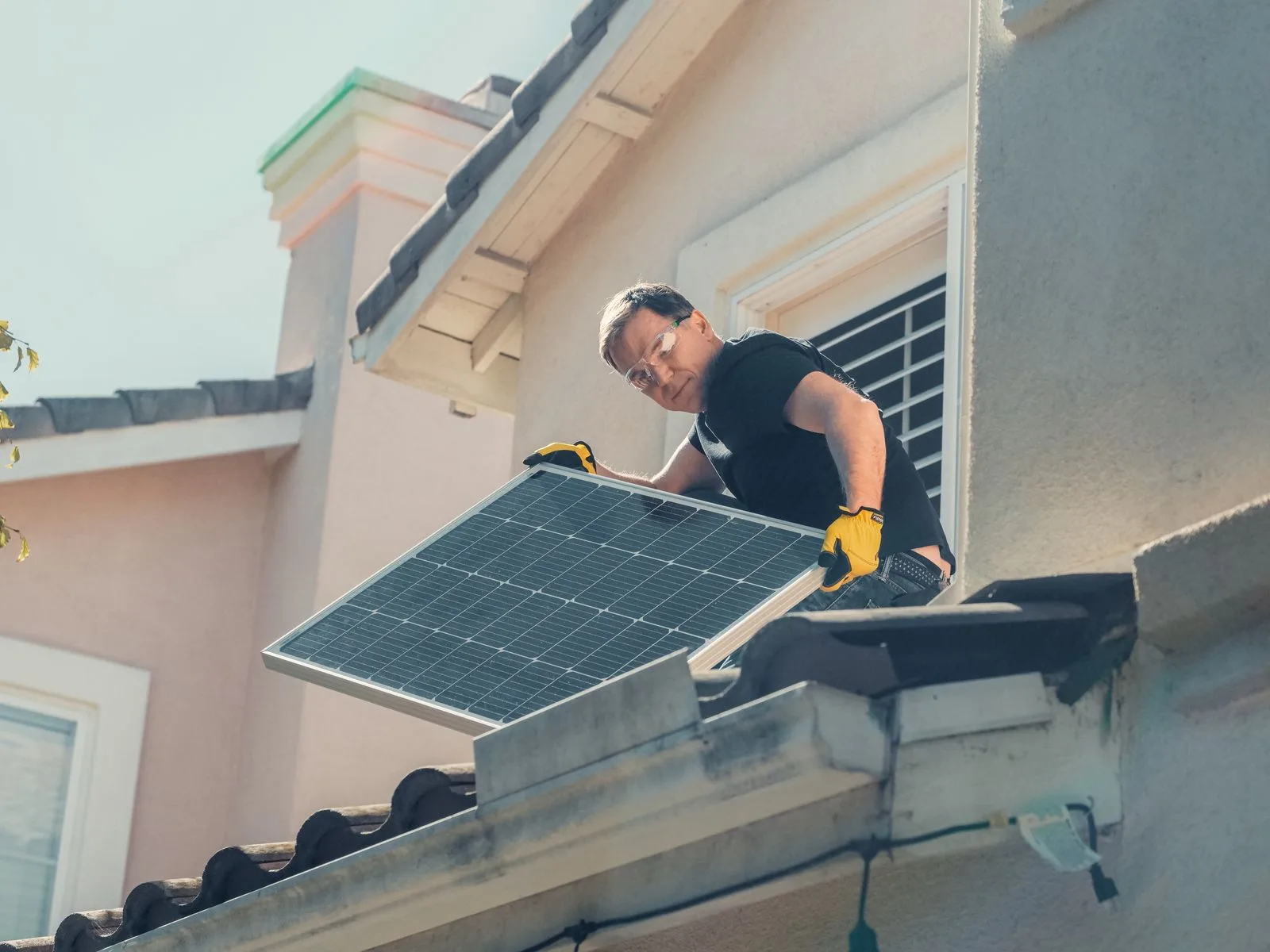 An installer in safety glasses lifting a solar panel onto a tiled roof